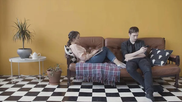 Jeune couple assis à la maison sur le canapé, accro et obsédé par les gadgets modernes. Images d'archives. Homme utilisant le téléphone mobile tandis que la petite amie surutilisant ordinateur portable, éviter de s'ignorer mutuellement . — Photo