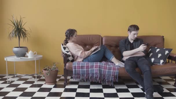 Молодая пара сидит дома на диване, зависимая и одержимая современными гаджетами. Запись. Человек, использующий мобильный телефон в то время как девушка злоупотребляет ноутбуком, избегая игнорировать друг друга . — стоковое видео