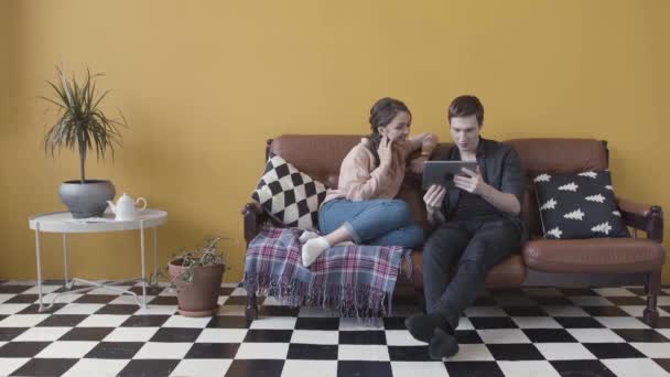 Couple sitting in sofa with electronic tablet in unusual room with yellow wall and black and white checkered floor. Stock footage. Cheerful couple at home e-shopping with tablet. — Stock Video