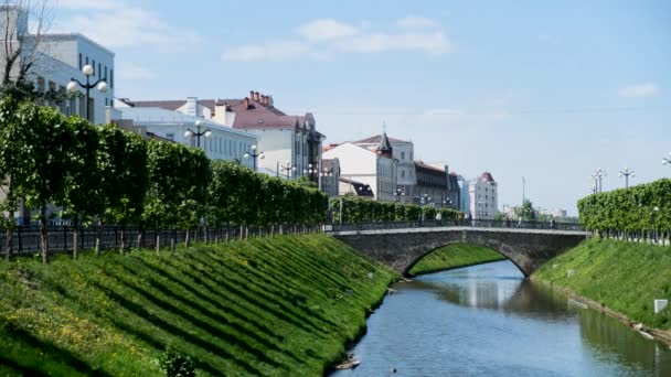 Paisaje urbano con un estrecho río rodeado de follaje verde y edificios blancos. Imágenes de archivo. Una pintoresca calle en verano con gente cruzando el puente . — Vídeo de stock