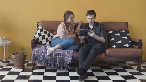 Joven pareja sonriente con tableta digital que se relaja en el sofá en casa en una habitación elegante. Imágenes de archivo. Hombre y mujer navegando en su dispositivo juntos . — Foto de Stock