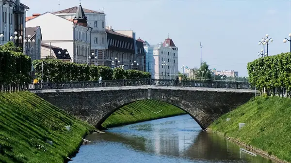 Cityscape s úzkou řekou obklopenou zeleným listím a bílými budovami. Záběry ze skladu. Malebná ulice v letním období s lidmi přecházejícími most. — Stock fotografie