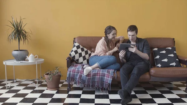 Paar sitzt auf Sofa mit elektronischem Tablet in ungewöhnlichem Raum mit gelber Wand und schwarz-weiß kariertem Boden. Archivmaterial. Fröhliches Paar zu Hause E-Shopping mit Tablet. — Stockfoto
