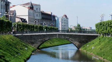 Dar bir kanalı ve mavi bulutlu arka planda yeşil çimlerle kaplı iki yamacı olan güzel bir şehir manzarası. Stok görüntüleri. Summer City Caddesi 'nde beyaz evler ve köprüde yürüyen insanlar..
