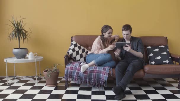 Pareja sentada en sofá con tableta electrónica en habitación inusual con pared amarilla y piso a cuadros en blanco y negro. Imágenes de archivo. Pareja alegre en casa e-shopping con tablet . — Vídeo de stock