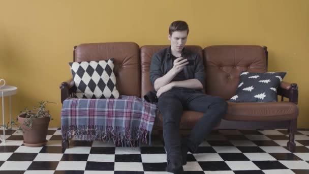 Ung kille sitter på den läderbruna soffan i vardagsrummet och tittar på något på sin mobiltelefon. Lagerbilder. Telefonberoende man som använder sin enhet. — Stockvideo