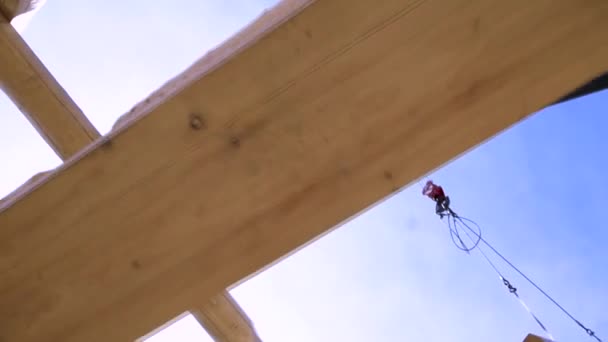 Construcción de casa de madera, grúa levantando un tronco de madera sobre fondo azul cielo nublado. Clip. Vista inferior del proceso de construcción de una nueva casa de madera . — Vídeo de stock