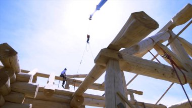 İnşaat alanındaki ahşap kirişin üzerinde oturan bir marangozun alt manzarası, iskelet. Şarjör. Ahşap kütük vinç tarafından taşınıyor..