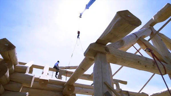 Blick von unten auf einen Tischler, der auf einem Holzbalken an der Baustelle sitzt, Gerüst. Clip. Holzstamm, der vom Kran getragen wird, Hausbau-Prozess. — Stockfoto