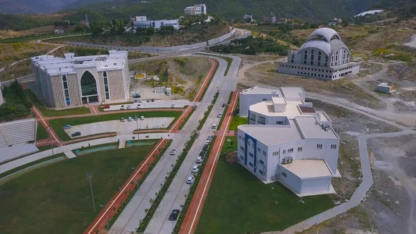 Aerial view of the univercity campus near the parking and green decorative lawn with pedestrian area. Clip. Top view of modern buildings on green forested hills background, education concept.