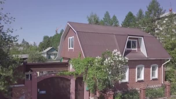 Zdjęcie uroczego domu z czerwonej cegły z kwitnącymi krzewami i zielonymi drzewami rosnącymi wokół niego. Materiał filmowy. Krajobraz z domami i liśćmi kołyszącymi się na wietrze. — Wideo stockowe
