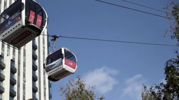 Cable cars on blue sky background with white clouds. Action. Bottom view of cabins moving slowly on a cable car road above green trees foliage. — Stockvideo