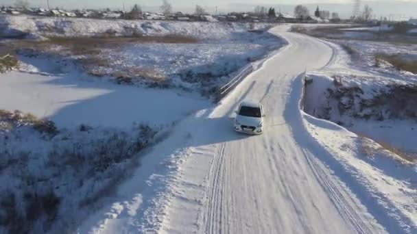 Ein weißes Auto überquert die schneebedeckte Brücke im Winterfeld, unter der ein schmaler zugefrorener Fluss fließt. Archivmaterial. Luftaufnahme eines fahrenden Autos auf einer winterlichen Straße mit einem Dorf dahinter. — Stockvideo