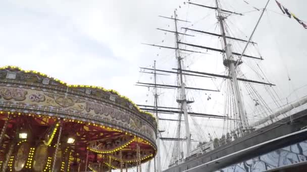 Prachtige carrousel op achtergrond van groot schip. Actie. Heldere carrousel spinnen op de achtergrond van zeilend havenschip bij bewolkt weer. Promenade met toeristische attracties — Stockvideo