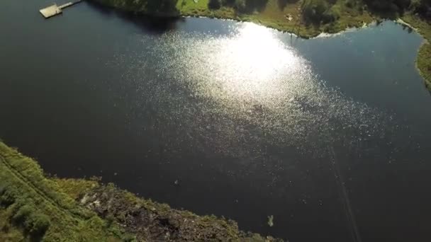 Повітряний літній пейзаж зеленої долини, маленьке село, і озеро з відображенням сонця на його поверхні. Стокові кадри. Вид зверху на зелену траву, будинки і озеро . — стокове відео