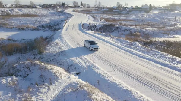 White car crossing the bridge covered with snow in the winter field with a narrow frozen river under it. Stock footage. Aerial view of a driving car on a winter road with a village behind.