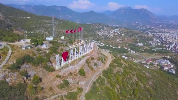 Turkse vlaggen op een rij en de inscriptie Ik hou van Alanya op de helling van een berg, toeristisch concept. Kunst. Prachtig land vanaf een hoogte met groene vegetatie en daken van gebouwen. — Stockvideo