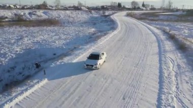 Kış tarlasında karla kaplı beyaz bir araba köprüyü geçiyor ve altında dar bir donmuş nehir var. Stok görüntüleri. Kış yolunda, arkada bir köy olan bir arabanın hava manzarası..