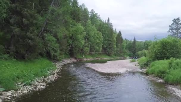 Picturesque forest river and old wooden bridge with group of people standing under it. Stock footage. Hikers standing under the suspension bridge on a stony shore near the river. — Stock Video