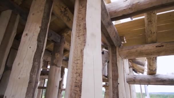 An employee in white uniform and glasses polishing the wooden wall of a cottage made of round logs. Clip. A carpenter working with a grinding machine at the construction site. — ストック動画