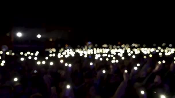 Espectadores desenfocados en la sala con luces ondeando en la oscuridad. Acción. Público sosteniendo teléfonos con linternas brillantes durante el concierto, ambiente romántico . — Vídeo de stock