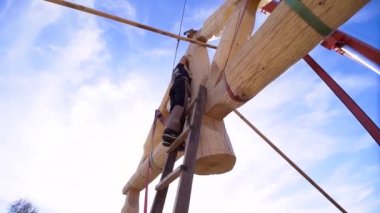Büyük bir evin çatısında çalışan bir marangozun arka görüntüsü. Şarjör. Mavi bulutlu arka planda merdivenlerin üzerinde duran bir inşaatçının alt görünümü.