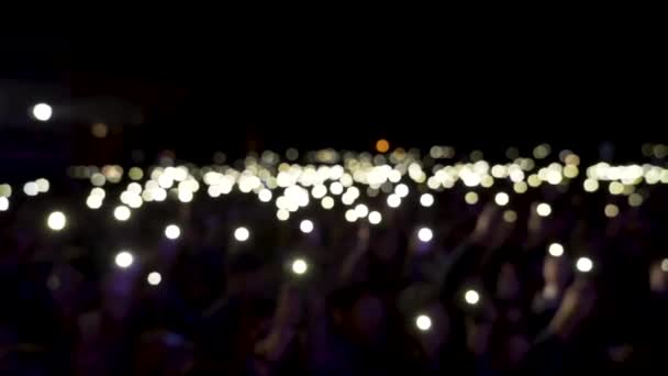 Espectadores desenfocados en la sala con luces ondeando en la oscuridad. Acción. Público sosteniendo teléfonos con linternas brillantes durante el concierto, ambiente romántico . — Vídeo de stock