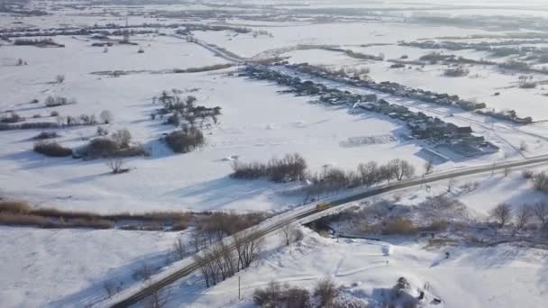 Landasfaltweg met gele bus in de winter en klein dorpje van boven. Neergeschoten. Luchtfoto drone zicht in koude winter sneeuw witte velden en bossen. — Stockvideo