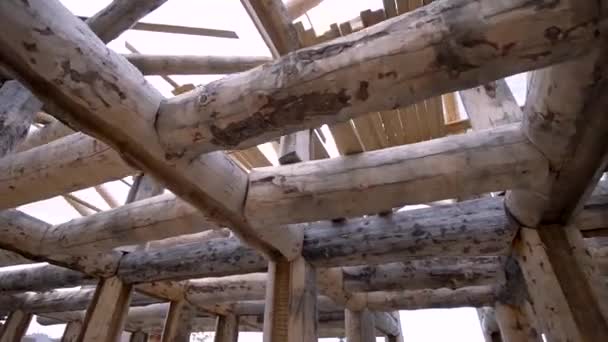 View of wooden log construction while installing roof on cloudy sky background. Clip. Bottom view of many beams inside the unfinished house. — ストック動画