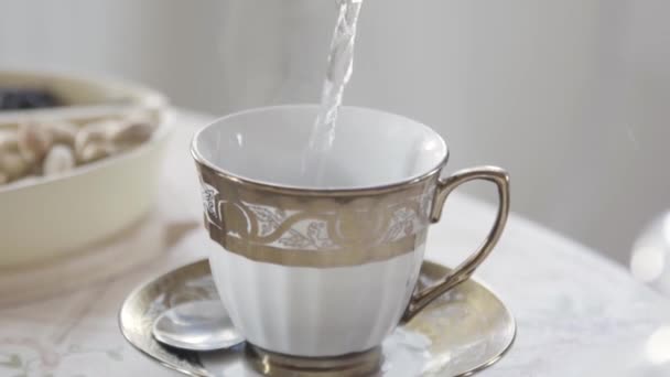 Közel a porcelán bögre fehér és arany színű áll az asztalon forró tea. Készletfelvétel. Forró vizet öntenek az asztalra egy kerámia pohárba.. — Stock videók