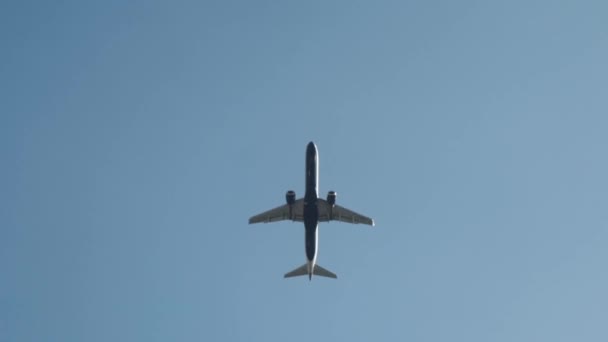 View from the bottom on a large white passenger plane flying against the clear blue sky. Action. Flight to the summer resort. — ストック動画