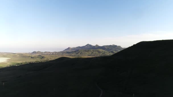 Aerial view of the small settlement in green hills with high mountain chain and blue sea on the background against blue sky. Shot. Beautiful colorful landscape. — Vídeos de Stock