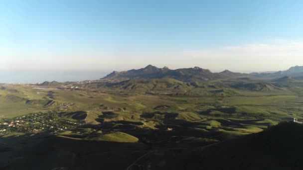 Aerial view of the small settlement in green hills with high mountain chain and blue sea on the background against blue sky. Shot. Beautiful colorful landscape. — Stock Video