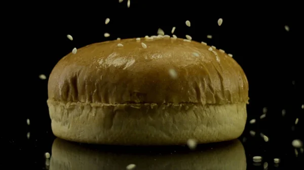 Close up de sementes de gergelim caindo no pão de hambúrguer branco isolado no fundo preto. Imagens de stock. Conceito de comida e cozinha ocidental . — Fotografia de Stock
