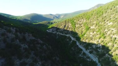 Aerial view of road going through a green mountains covered with shrubs and grass against blue sky in subber day. Shot. Beautiful view from above
