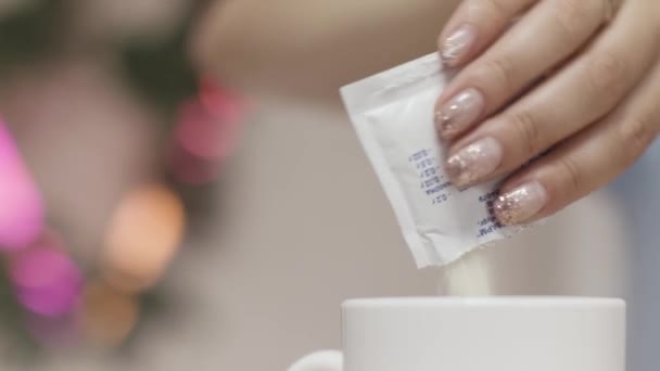 Close up of a woman hands throwing sugar into a white mug with hot beverage on blurred background. Stock footage. Female hands adding white sugar into tea or coffee. — 图库视频影像