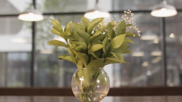 Gentle lilly of valley flowers in glass vase standing on wooden table on blurred background with windows and lamps. Stock footage. Spring may lily bouquet, decoration of room. — 비디오