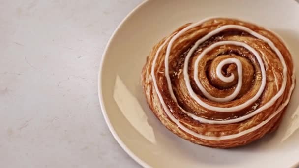 Close up top view of real sweet and fresh bun with white glaze lying on the plate on white table background. Stock footage. Perfect breakfast, foodporn concept. — Stock Video