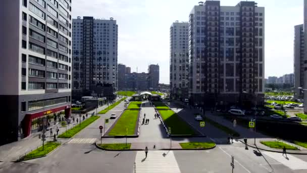 Uzun modern evleri olan şehir merkezi, yaya geçidinde yürüyen insanlar ve güneşli bir yaz gününde yeşil bir sokak. Hareket. Yeşil çimlerle süslenmiş güzel caddenin havadan görünüşü. — Stok video