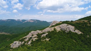 Mavi gökyüzünün arka planında bulutlu dağların olduğu tepelik bölgenin havadan görünüşü. Vuruldu. Dağların üzerinden uçarken sık yeşil orman ve kaya oluşumları örtüldü..