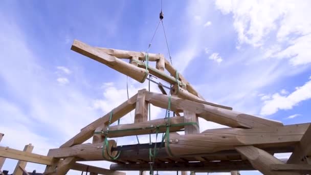 Kran über dem im Bau befindlichen Holzgebäude an einem sonnigen Tag im Grünen. Clip. Holzbalkenkonstruktion wird über den Hausrahmen transportiert. — Stockvideo