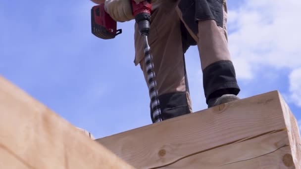 木造フレームを組み立て、家を建てるときにログに穴を作るための金属スパイラルドリル。クリップ。木製の梁の内側に穴を掘る男の下のビュー. — ストック動画
