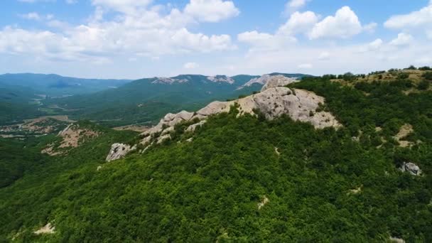 Luftaufnahme einer hügeligen Region mit Bergen vor blauem Himmel mit Wolken. Schuss. fliegen über die mit dichten grünen Wäldern und Felsformationen bedeckten Berge. — Stockvideo