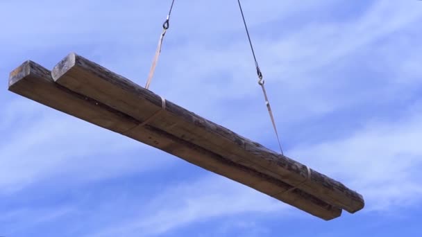 Twee houten balken worden door de kraan op een blauwe wolkenbasis getransporteerd. Een knip. Onderaanzicht van de logs in de lucht. — Stockvideo
