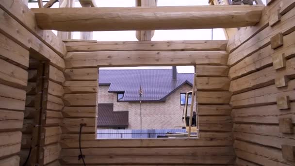 Vista de dentro de uma nova casa em construção feita de troncos de madeira com uma máquina tratora fora da janela. Clipe. Conceito de obras de carpintaria . — Vídeo de Stock