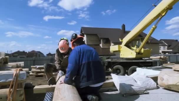 La vista trasera de los carpinteros hace marcas en un tronco de madera en el sitio de construcción con la máquina de grúa en el fondo. Clip. Proceso de construcción de casa de madera nueva . — Vídeo de stock