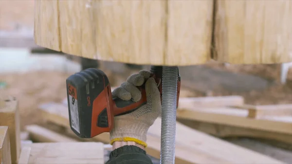 De arbeider werkt met een schroevendraaier, op de achtergrond van houten balken, timmerwerk op de bouwplaats. Een knip. Close-up van mannelijke hand losschroeven metalen bout uit houten ronde log. — Stockfoto