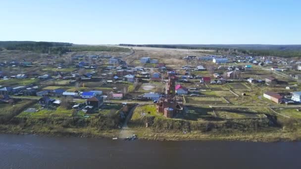 Flygfoto av gamla förstörda kyrkan ligger vid floden stranden nära den lilla byn. Klipp. Övergivna gamla ruiner av kapellet och hus på blå molnig himmel bakgrund. — Stockvideo