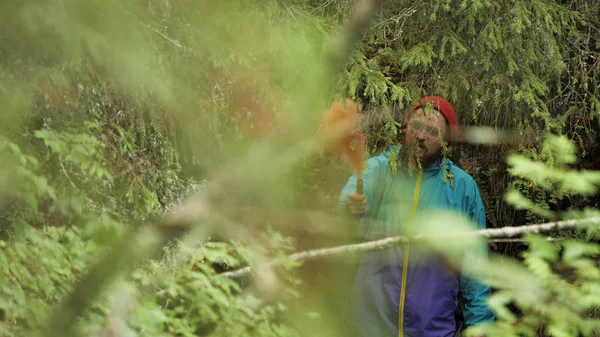 Hombre de pie en el bosque verde matorral y sosteniendo una llamarada de humo brillante en su mano. Imágenes de archivo. Viajero con chaqueta azul y sombrero rojo se perdió en el bosque . — Foto de Stock