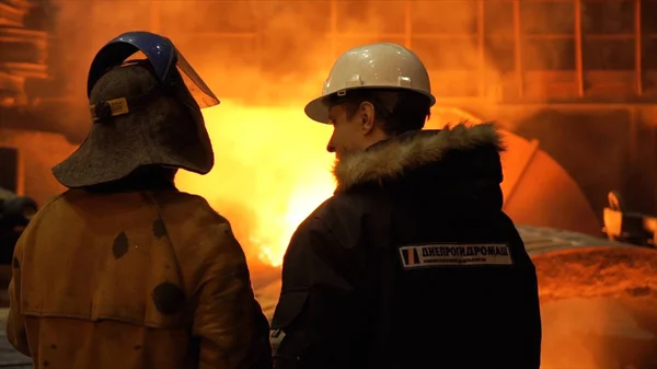 Rückansicht eines Arbeiters in feuerfester Uniform und eines Ingenieurs, der vor einem funktionierenden Hochofen in der Fabrik steht. Archivmaterial. Blick ins Innere der Hüttenmanufaktur. — Stockfoto